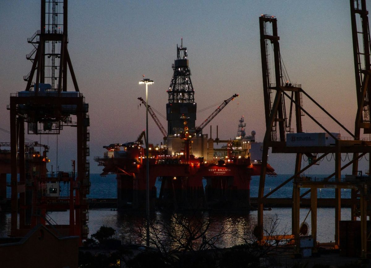 Offshore drilling rig lit at dusk alongside quayside cranes